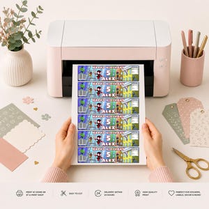 May include: A printed sheet of rectangular labels featuring a superhero theme with the text Happy Birthday 5 Alex. The labels include colorful character graphics, a barcode, and a city background. The sheet is held in front of a white desktop printer.
