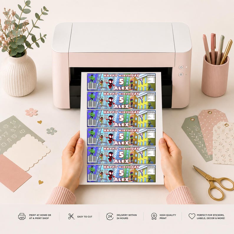 May include: A printed sheet of rectangular labels featuring a superhero theme with the text Happy Birthday 5 Alex. The labels include colorful character graphics, a barcode, and a city background. The sheet is held in front of a white desktop printer.