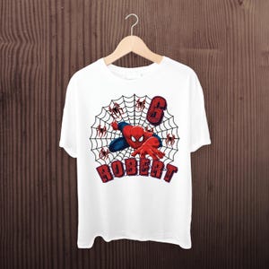 May include: A white t-shirt featuring a graphic of a red and blue superhero character crawling on a black spider web. The design includes the number 6 in a red textured font and the name ROBERT in red block letters with a dark outline.