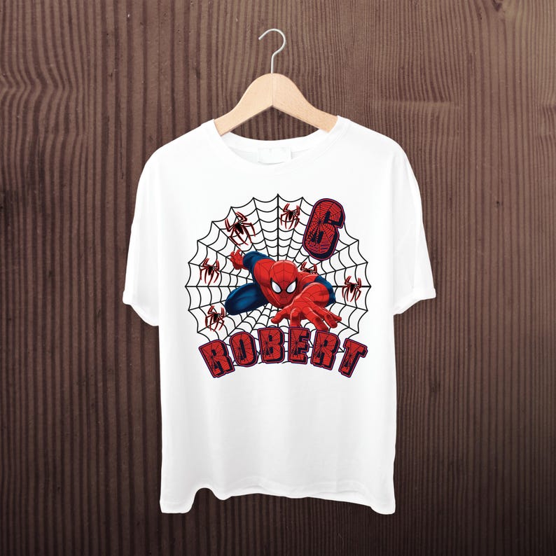 May include: A white t-shirt featuring a graphic of a red and blue superhero character crawling on a black spider web. The design includes the number 6 in a red textured font and the name ROBERT in red block letters with a dark outline.