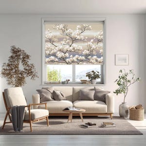 May include: A window roller blind featuring a detailed print of white magnolia blossoms on dark branches against a soft beige and light blue gradient background. The blind is installed inside a window frame above a beige sofa and armchair.