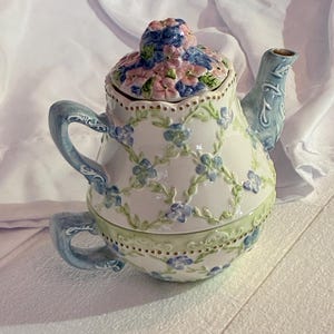 May include: A ceramic teapot for one featuring a stacked cup design. The white surface is decorated with a raised green vine trellis pattern and small blue flowers. It includes a matching lid with sculpted floral details and a blue handle.