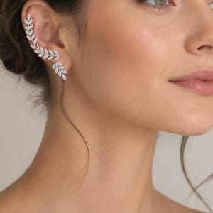 Op de afbeelding: Een zilverkleurige ear crawler oorbbel met een delicaat blad- of rankmotief, ingelegd met kleine, heldere, fonkelende steentjes. Het sieraad volgt de natuurlijke welving van het oorkraakbeen en loopt vanaf de oorlel omhoog langs de rand.