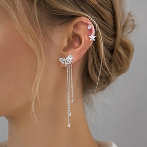 May include: A silver-toned ear cuff and earring set featuring clear, sparkling stones. The ear cuff displays two small floral-shaped accents, while the earring showcases a leaf-like cluster with three delicate dangling chains tipped with stones.
