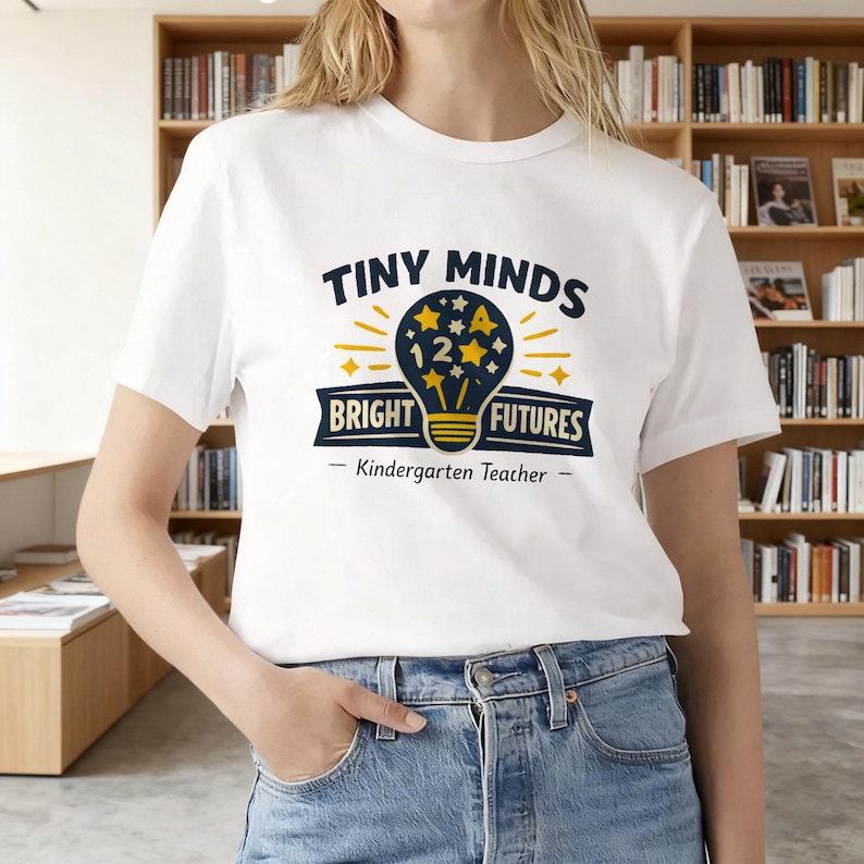 May include: A white short-sleeve t-shirt featuring a graphic of a dark blue lightbulb with yellow stars and the number 12 inside. The text reads "TINY MINDS BRIGHT FUTURES" and "Kindergarten Teacher" in dark blue and yellow lettering.
