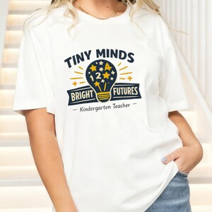 May include: A white short-sleeve t-shirt featuring a graphic of a lightbulb filled with stars and the number 12. The text reads "TINY MINDS BRIGHT FUTURES" and "Kindergarten Teacher" in dark blue and yellow ink on the front of the garment.