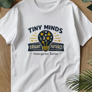 May include: A white short-sleeved t-shirt featuring a graphic of a dark blue lightbulb filled with yellow stars and the number 12. The text reads "TINY MINDS BRIGHT FUTURES" and "Kindergarten Teacher" in dark blue and yellow lettering.