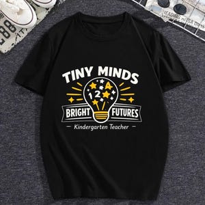 May include: A black short-sleeved t-shirt featuring a graphic print with the text "TINY MINDS BRIGHT FUTURES" and "Kindergarten Teacher". The design includes a yellow lightbulb icon containing the number 12 and several small yellow stars.