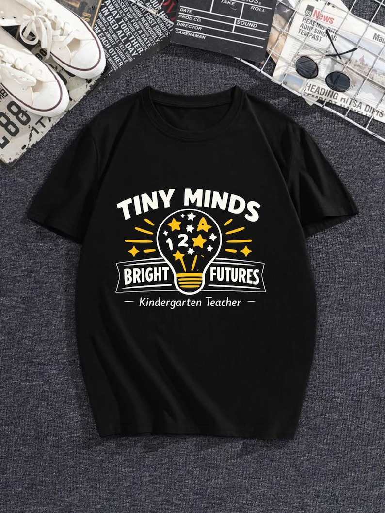 May include: A black short-sleeved t-shirt featuring a graphic print with the text "TINY MINDS BRIGHT FUTURES" and "Kindergarten Teacher". The design includes a yellow lightbulb icon containing the number 12 and several small yellow stars.