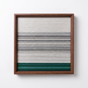 Puede incluir: Una pieza de arte mural cuadrada que presenta capas horizontales de hilo enrollado apretadamente en tonos blanco, gris claro, gris medio y gris oscuro, terminada con una banda sólida de verde bosque profundo en la parte inferior, enmarcada en madera marrón.
