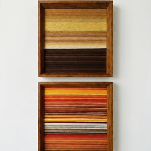 May include: Two square wall art pieces featuring horizontal yarn layers in wooden frames. The top piece displays yellow, tan, and dark brown gradients. The bottom piece features a vibrant pattern of red, orange, yellow, and grey horizontal stripes.