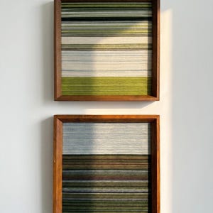 May include: Two square wall art pieces featuring horizontal yarn strands stretched across wooden frames. The top piece displays shades of green, white, and grey. The bottom piece features horizontal bands of dark brown, grey, and olive green yarn.