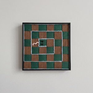 May include: A square wall art piece featuring a woven checkerboard pattern in dark green and brown yarn. A light blue yarn outline forms a spiral path across the surface, containing two small black eyes and a pink wavy yarn tongue at the center.