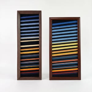 May include: Two rectangular wall art pieces in dark wood frames. Each frame contains a series of horizontal, angled stripes in shades of blue, yellow, orange, and red set against a black background, creating a layered, textured appearance.