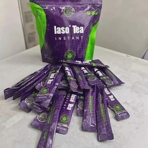May include: A large purple and green pouch of Iaso Tea Instant with a pile of matching individual tea stick packets spread out in front. The packaging features the brand name Iaso Tea in white text, along with circular green logos and patterns.