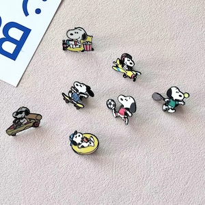 May include: A collection of seven enamel pins featuring the character Snoopy in various activities, including sitting with popcorn, flying a yellow airplane, skateboarding, playing soccer, playing tennis, driving a race car, and floating in a pool ring.