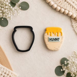 Può includere: Un tagliabiscotti in plastica nera a forma di vasetto di miele accanto a un biscotto decorato a forma di vasetto di miele. Il biscotto presenta una glassa gialla che cola dall'alto, una base color sabbia e un'etichetta con la scritta HUNNY.