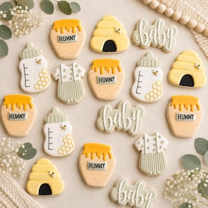 Puede incluir: Una colección de galletas decoradas con tarros de miel etiquetados como HUNNY, colmenas amarillas, biberones con patrones de panal, bodies blancos con estampados de abejas y galletas con la palabra baby escrita en glaseado blanco.