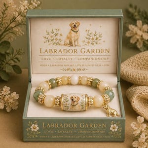 May include: A beaded bracelet with a central cylindrical charm featuring a yellow Labrador illustration, flanked by green and cream beads and gold-tone spacers. A small Labrador charm hangs from the bracelet, presented in a green box with text.