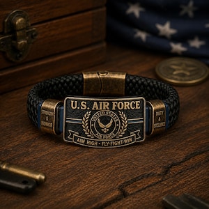 May include: A black braided leather bracelet featuring a rectangular antique brass-toned metal plate with the text U.S. AIR FORCE, the Air Force emblem, and the motto AIM HIGH FLY-FIGHT-WIN. Side plates are engraved with STRENGTH & HONOR and DUTY & EXCELLENCE.