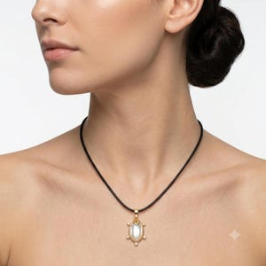 May include: A gold-toned pendant shaped like a turtle with a smooth, oval-shaped white stone center, suspended from a thin black cord necklace. The pendant features small gold-toned accents on the head, legs, and tail, creating a detailed turtle design.