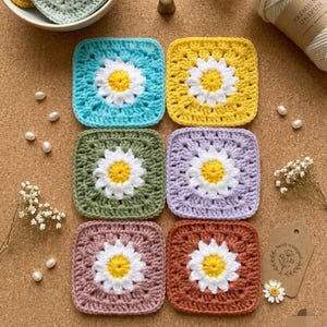 May include: Six square crochet motifs arranged in two columns, each featuring a central white and yellow daisy pattern. The squares are colored in light blue, yellow, sage green, lavender, dusty rose, and terracotta, crafted with yarn.
