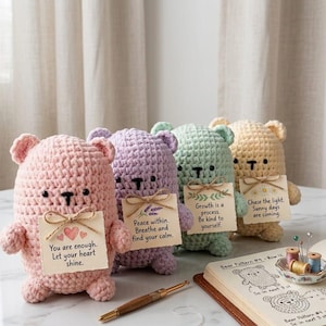 May include: Four crocheted plush bears in pastel pink, lavender, mint green, and cream. Each bear holds a square card with inspirational text tied with twine. A notebook with handwritten crochet patterns and a gold pen sit in the foreground.