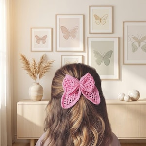 May include: A bright pink crochet hair accessory shaped like a butterfly with an openwork, textured stitch pattern. The accessory is secured in long, wavy brown hair, featuring a layered design with two larger upper wings and two smaller lower wings.
