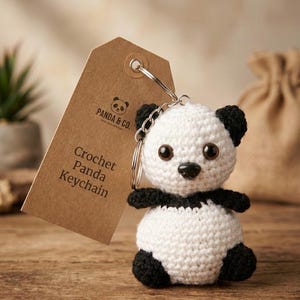 May include: A small, round, handmade crochet panda keychain with black and white yarn, featuring black plastic eyes and a black nose. Attached to the panda is a silver metal keyring and a brown cardboard tag printed with "PANDA & CO. Crochet Panda Keychain".