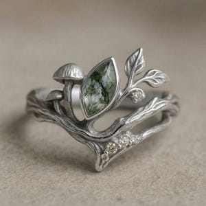 May include: A silver ring featuring a marquise-cut moss agate stone, two small mushroom sculptures, and delicate leaf motifs. The band is textured to resemble a tree branch and is accented with several tiny, clear round-cut gemstones.