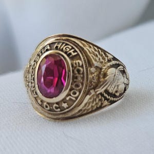 May include: A gold-coloured Anacostia High School class ring with a large, oval, ruby-red stone. The ring features the school's name and "D.C." engraved around the stone. Intricate details and a Native American profile are on the ring's sides.