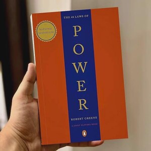 May include: A paperback book titled The 48 Laws of Power by Robert Greene and Joost Elffers. The cover features a vertical blue stripe on an orange background with gold text. A gold circular seal on the top left reads National Bestseller.