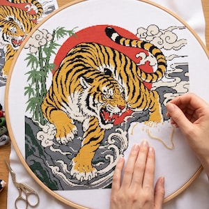 May include: A cross-stitch project in a round wooden embroidery hoop featuring a tiger with yellow and black stripes, a red sun background, green bamboo stalks, and stylized white clouds. The design is stitched on white fabric with a needle.