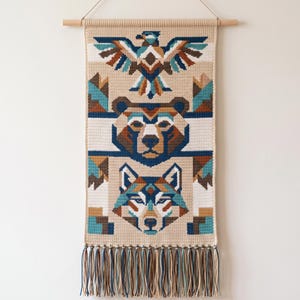 Puede incluir: Un tapiz de ganchillo rectangular suspendido de un listón de madera, con un patrón geométrico de un águila, un oso y un lobo en tonos azul, marrón y crema. La pieza está rematada con un fleco de borlas de hilo de varios colores.