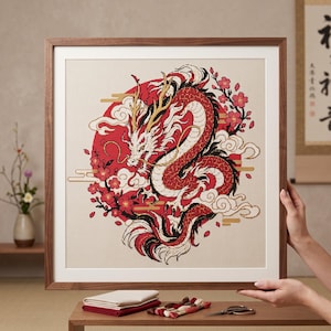 May include: A framed cross-stitch embroidery featuring a detailed red and white dragon coiled in front of a large red circle. The design includes stylized white clouds and pink cherry blossom branches on a light beige fabric background.