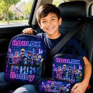 May include: A dark blue backpack, lunch bag, and pencil case set featuring a colorful Roblox graphic with various character figures and the name Bobby printed in neon pink text. Each item has a matching design with a dark background and neon accents.