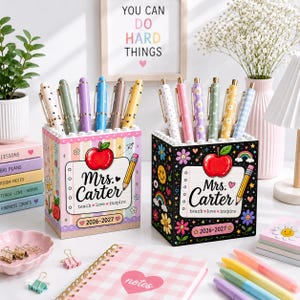 May include: Two square pencil holders featuring a red apple graphic, the text Mrs. Carter, teach love inspire, and 2026-2027. One holder has a pastel striped pattern, the other is black with colorful flowers, rainbows, and smiley faces.