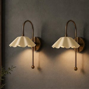 May include: A pair of wall sconces featuring a curved bronze-toned metal arm and a cream-coloured, scalloped-edge shade. Each lamp is mounted to a circular base and emits a soft, downward-facing light, creating a warm glow against the wall surface.