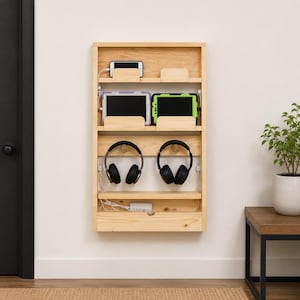 Peut inclure: Une station de charge murale en bois clair dotée de trois étagères horizontales. L'étagère supérieure accueille un smartphone, celle du milieu deux tablettes, et la section inférieure comprend deux crochets pour casques et une multiprise.