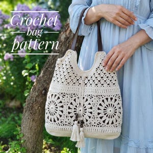 May include: A cream-colored crochet bag featuring intricate circular floral motifs and a drawstring closure with two dangling tassels. The bag has dark brown leather-like straps and is displayed against a natural outdoor background with text.
