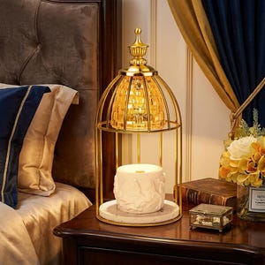 May include: A gold-toned metal birdcage-style candle warmer lamp featuring a decorative finial top and a faceted amber glass shade. The base holds a white cylindrical ceramic wax melt warmer with embossed horse figures on a circular marble tray.