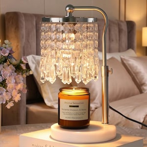 May include: A metal candle warmer lamp with a brushed silver finish, featuring a cylindrical shade adorned with hanging clear crystal prisms. The lamp sits on a white circular base, illuminating a brown glass jar candle labeled Aroma Sensation.