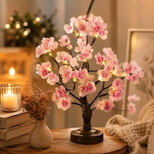 May include: A decorative LED tree lamp featuring dark brown branches adorned with numerous light pink cherry blossom flowers. The tree is illuminated by small warm white lights nestled among the petals, supported by a round black base.