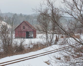 May include: A red barn with a white door and windows sits in a snowy field with a set of train tracks in the foreground. The barn is surrounded by bare trees and the ground is covered in snow.