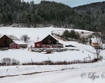 May include: A snowy winter scene with a red barn and other buildings on a snowy field. The barn has a wreath on the door and a stone wall in front of it. There are trees in the background.