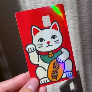 May include: A rectangular card with a bright red holographic finish featuring a white beckoning cat illustration. The cat wears a green collar with a bell and holds an oval gold coin with black Chinese characters. A small square cutout is at the top.