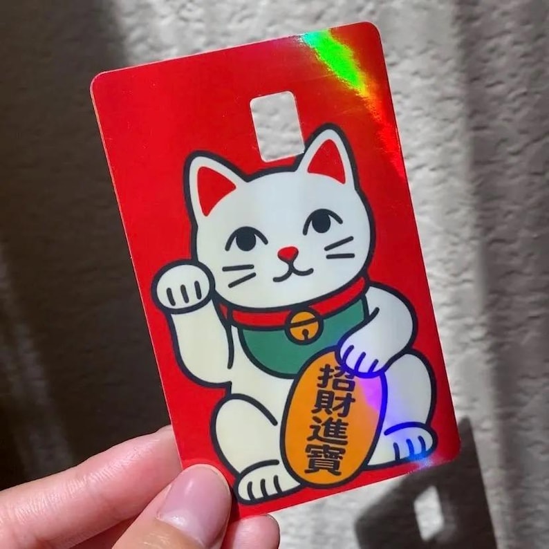 May include: A rectangular card with a bright red holographic finish featuring a white beckoning cat illustration. The cat wears a green collar with a bell and holds an oval gold coin with black Chinese characters. A small square cutout is at the top.
