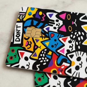 May include: A rectangular credit card skin featuring a vibrant, multi-colored cartoon pattern of various cats in black, white, yellow, blue, green, and red. The design includes the text DON'T printed in black on a white rectangular background.