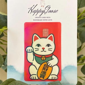 May include: A rectangular credit card skin featuring a white beckoning cat with a green collar and a gold coin with black Chinese characters. The background is a vibrant red with a subtle iridescent sheen. The skin is attached to a white display card.