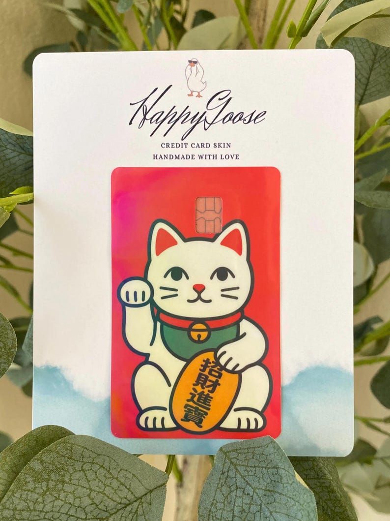 May include: A rectangular credit card skin featuring a white beckoning cat with a green collar and a gold coin with black Chinese characters. The background is a vibrant red with a subtle iridescent sheen. The skin is attached to a white display card.
